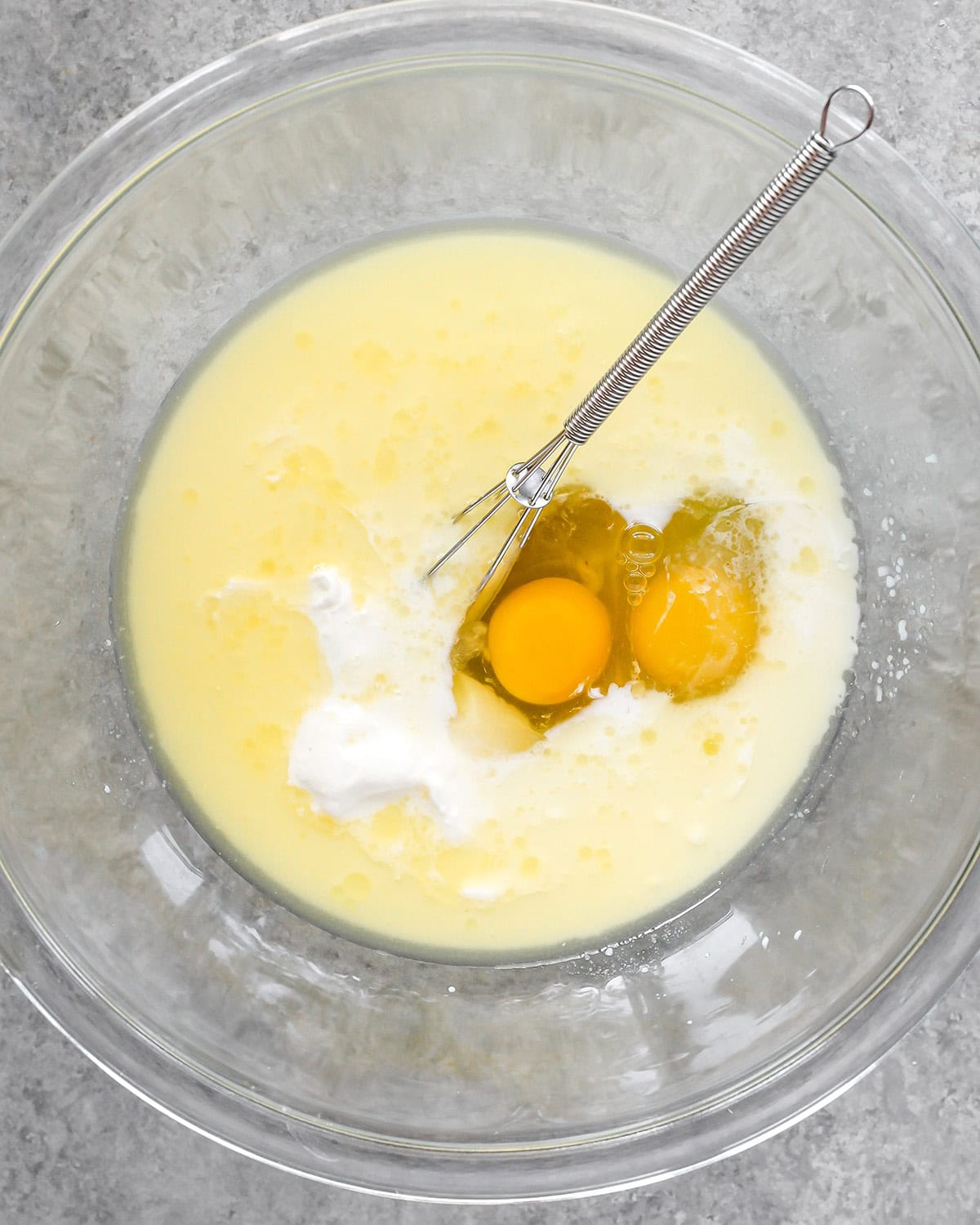 photo showing how to make cornbread - eggs, milk and sour cream added to the wet ingredients in a glass bowl with a metal wire whisk after mixing
