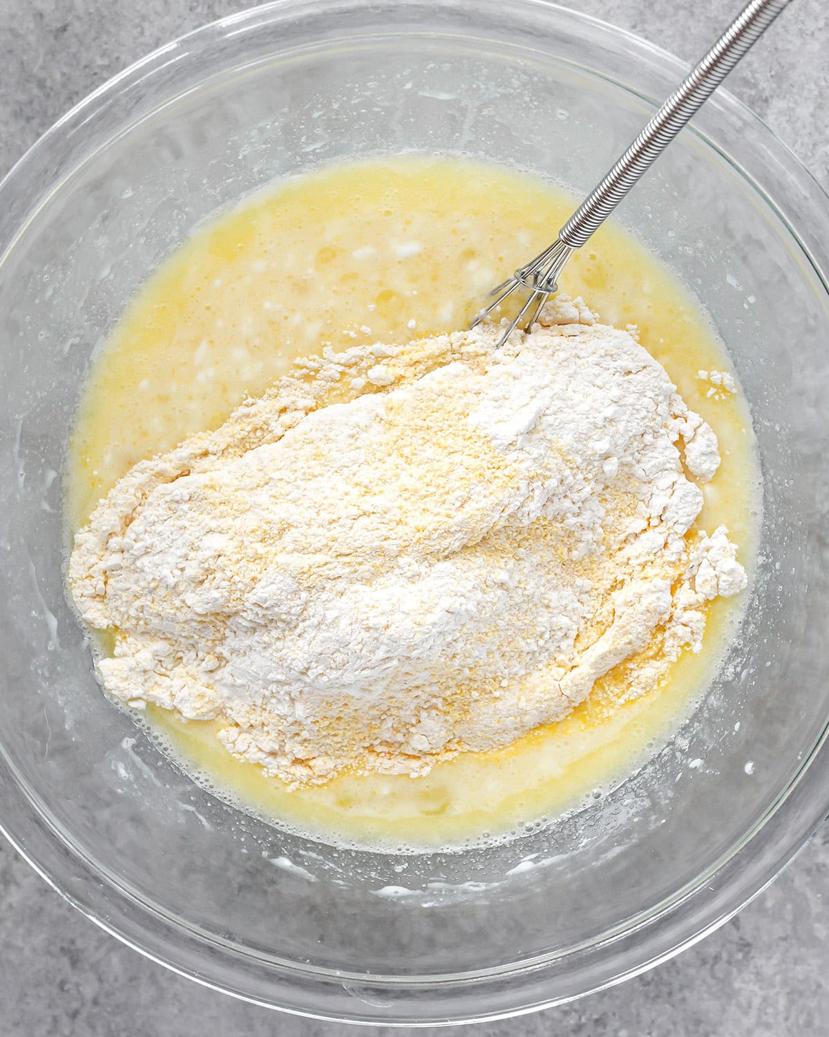 photo showing how to make cornbread - dry ingredient mixture added to the wet ingredient mixture in a glass bowl on a gray surface with a metal wire whisk int he bowl, before mixing