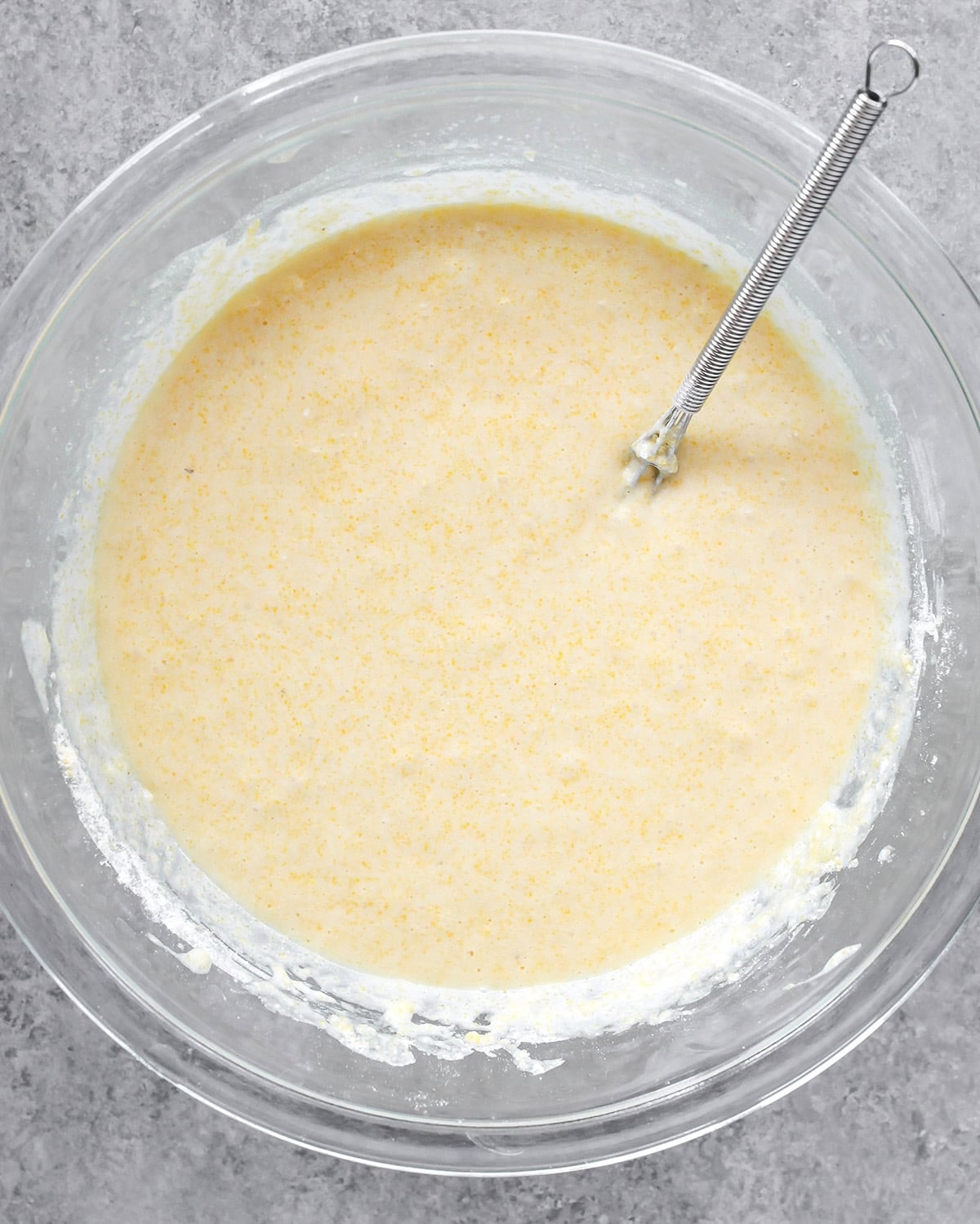 photo showing how to make cornbread - batter in a glass bowl with a wire metal whisk on a gray surface
