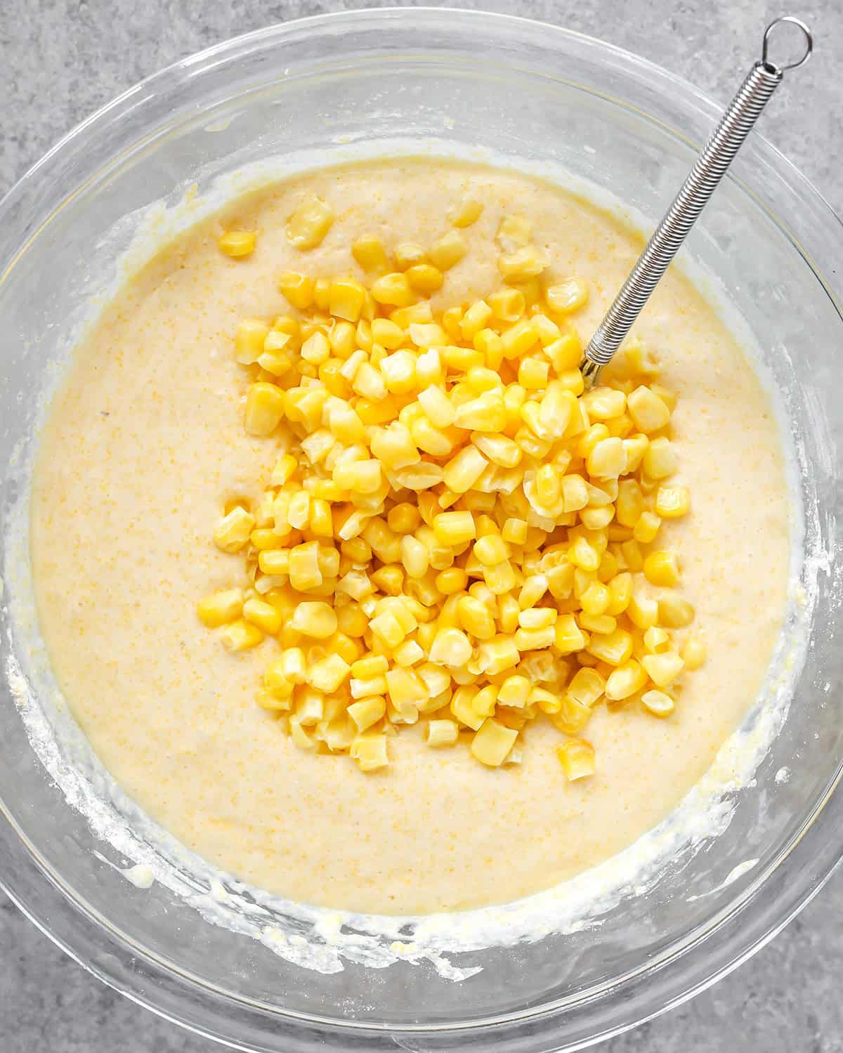 photo showing how to make cornbread - corn kernels added to the batter in a glass bowl on a gray surface with a metal whisk in th ebowl