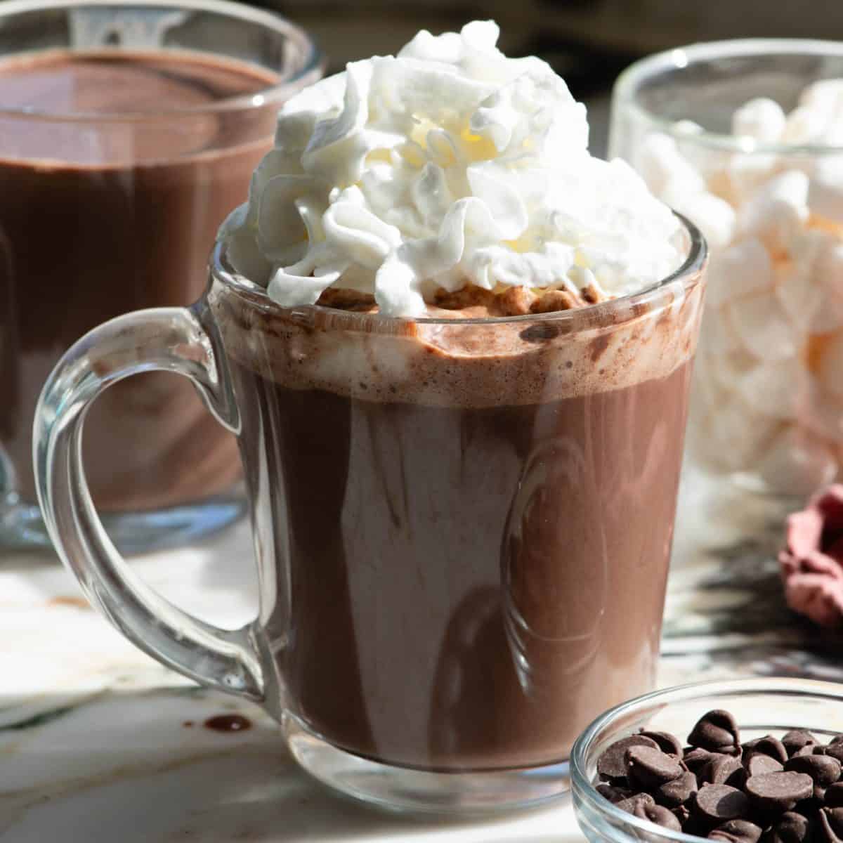 close up view of a glass mug filled with homemade hot chocolate recipe topped with fluffy whipped cream, there's another mug of hot chocolate in the background and a bowl of chocolate chips in the bottom right corner