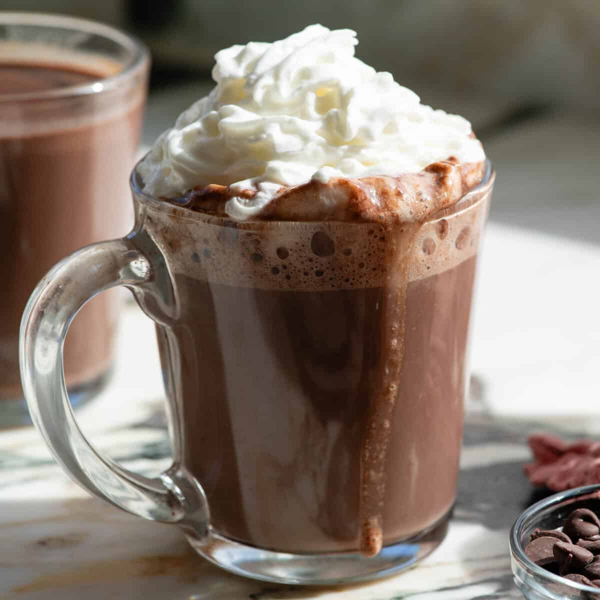 The best homemade hot chocolate recipe. This hot cocoa is smooth, creamy and made with two forms of chocolate for a rich, deep chocolate flavor. It’s easy to make in 5 minutes with 5 ingredients. front view of a glass mug filled with homemade hot chocolate recipe topped with fluffy whipped cream and a drip of hot chocolate going down the side of the mug, there's another mug of hot chocolate in the background and a bowl of chocolate chips in the bottom right corner