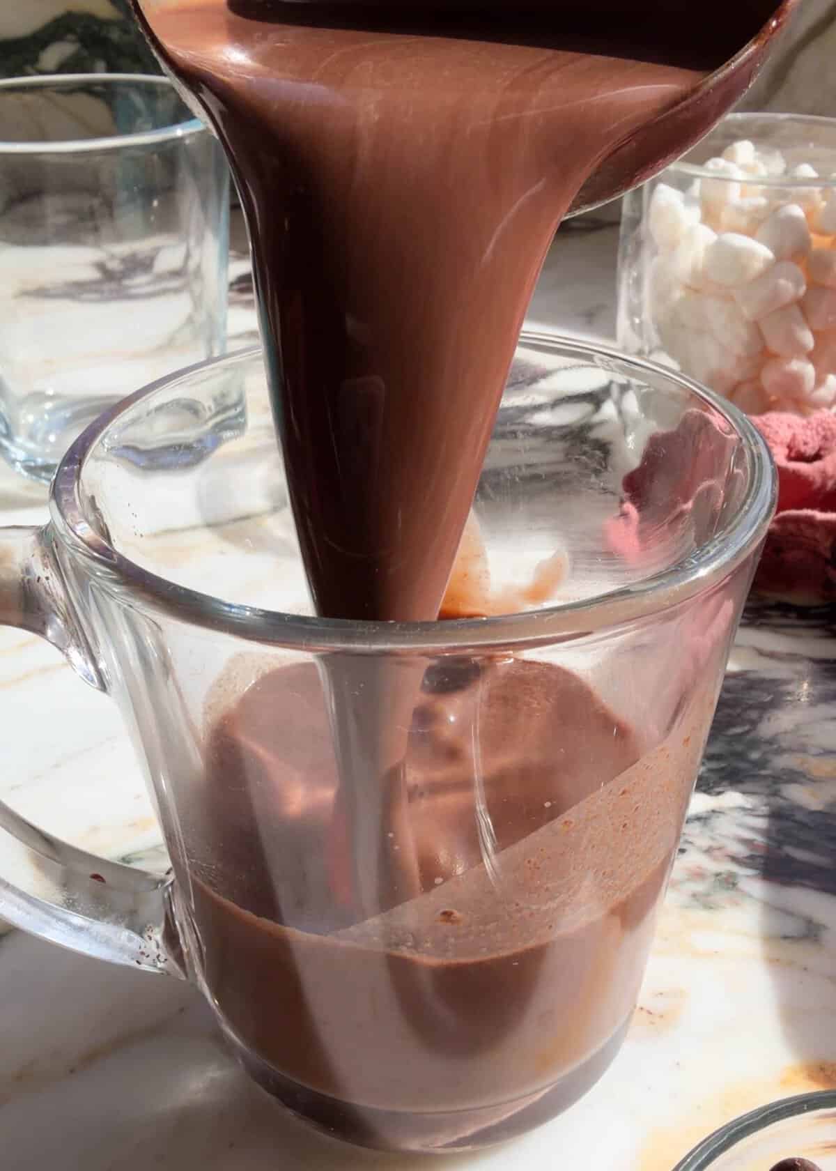 The best homemade hot chocolate recipe. This hot cocoa is smooth, creamy and made with two forms of chocolate for a rich, deep chocolate flavor. It’s easy to make in 5 minutes with 5 ingredients. homemade hot chocolate. recipe being poured into a glass mug from a metal ladle so the silky-smooth, rich chocolatey texture is visible. There's another empty glass mug in the back left corner and a glass mug filled with marshmallow in the back right corner.