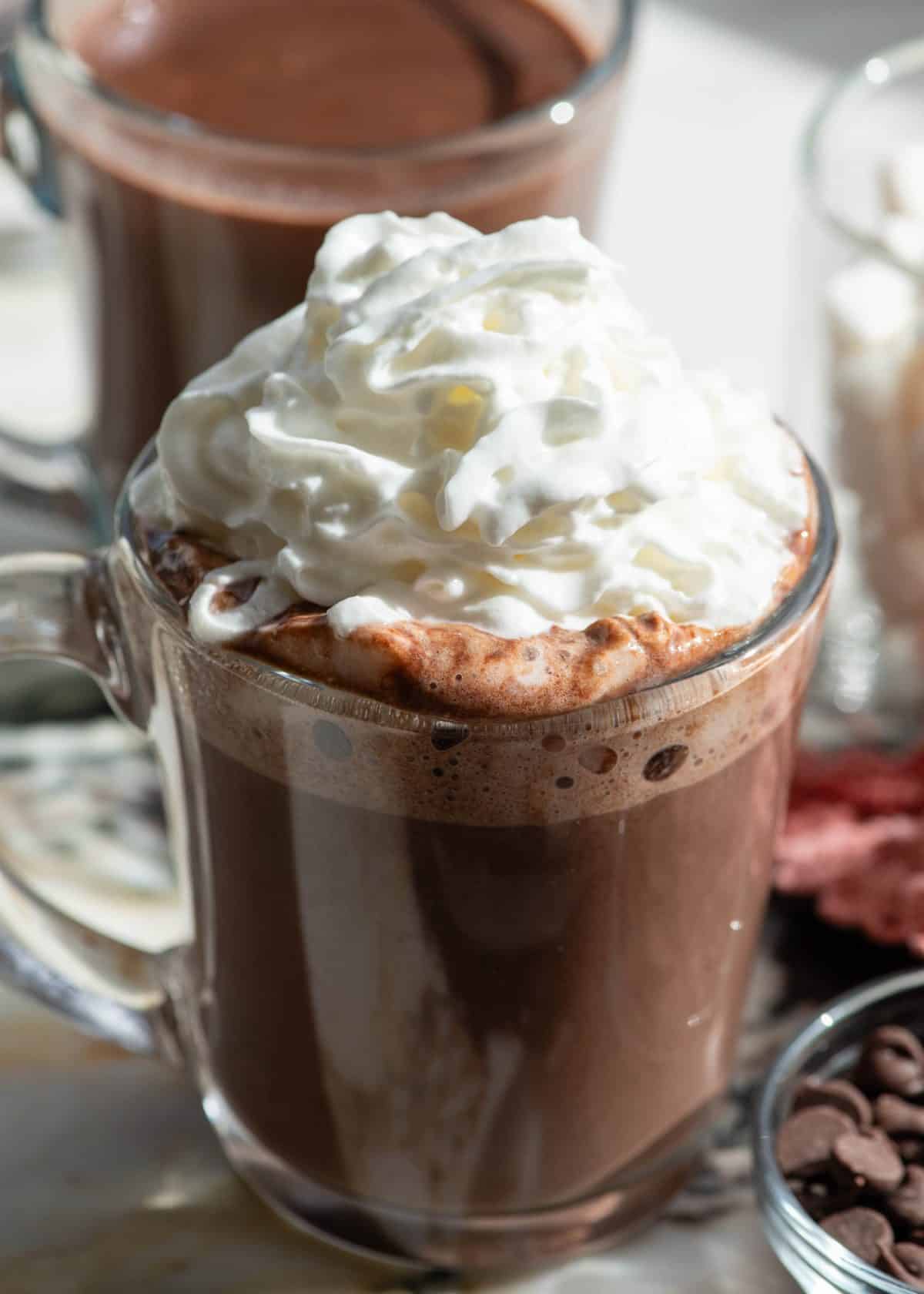 close up view of a glass mug filled with homemade hot chocolate recipe topped with fluffy whipped cream, there's another mug of hot chocolate in the background and a bowl of chocolate chips in the bottom right corner
