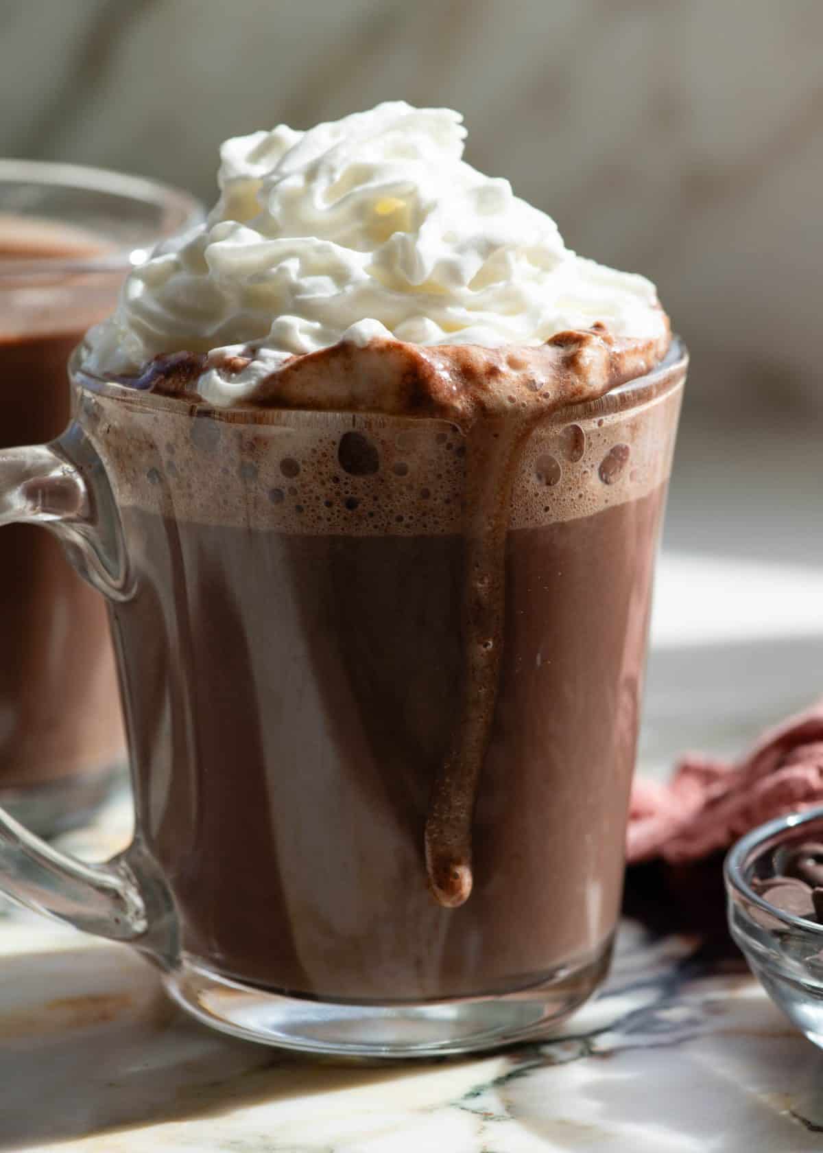 front view of a glass mug filled with homemade hot chocolate recipe topped with fluffy whipped cream and a drip of hot chocolate going down the side of the mug, there's another mug of hot chocolate in the background and a bowl of chocolate chips in the bottom right corner