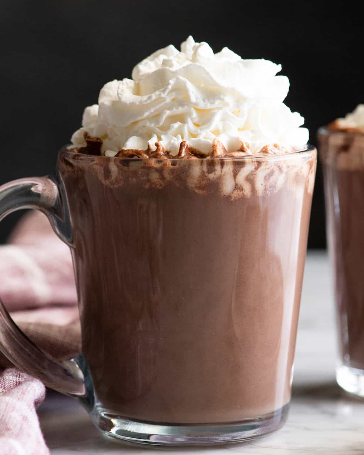 front view of a glass mug filled with homemade hot chocolate recipe topped with fluffy whipped cream, there's another mug and a towel in the background.