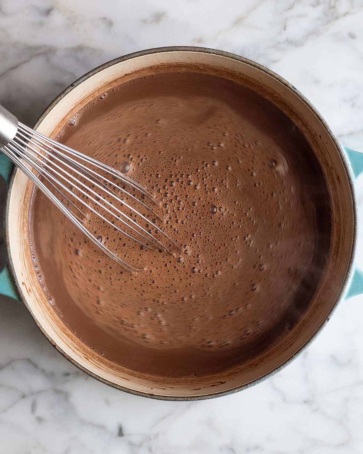 making homemade hot chocolate recipe - all the ingredients in a blue dutch oven being whisked together by a metal whisk