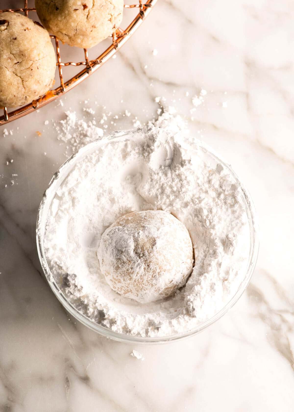 Pecan Snowball Cookies are easy to make with 7 ingredients. They are buttery, nutty, and melt-in your mouth delicious. These butterball cookies are an old family recipe, and a must-make Christmas cookie! one snowball cookie in a glass bowl filled with powdered sugar being coated with it, there's a copper wire cooing rack with uncoated snowball cookies in the top left corner.
