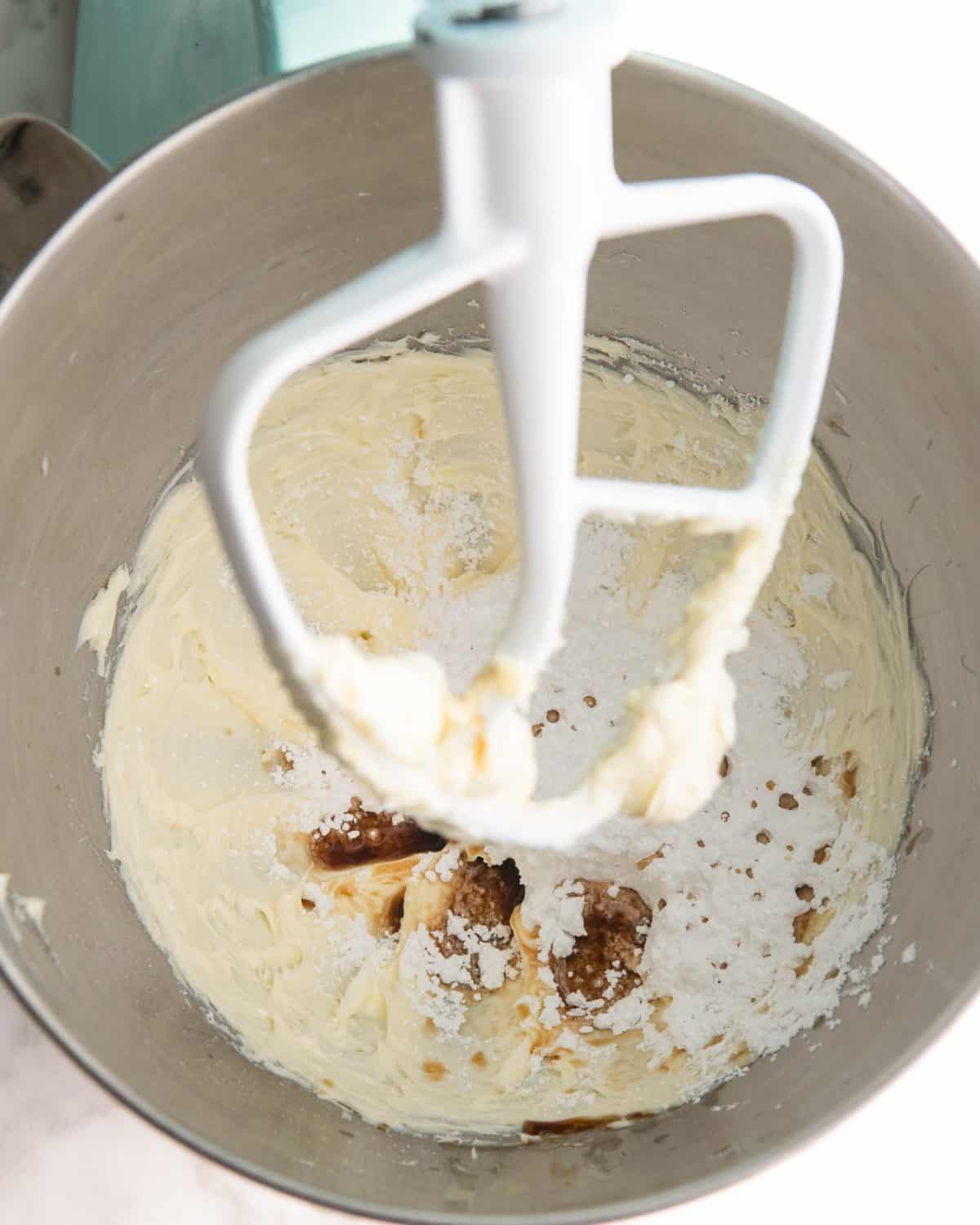 making pecan snowball cookies recipe - powdered sugar and vanilla added to the butter beaten in a light blue kitchen aid fitted with a metal mixing bowl and white paddle attachment