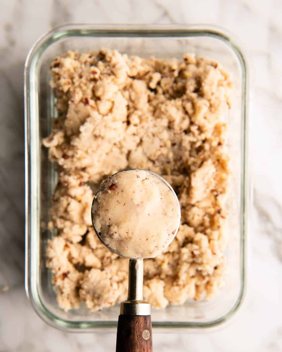 the dough for this pecan snowball cookies recipe in a rectangular glass container on a marble surface, theres a 1 Tablespoon measuring spoon filled with dough being held over the glass container of dough