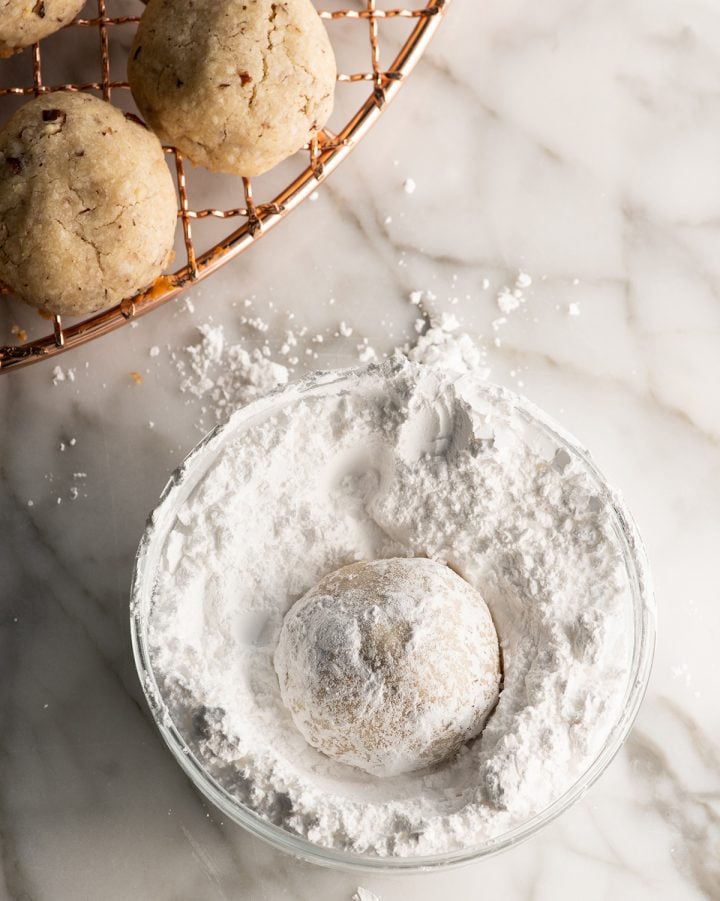 Pecan Snowball Cookies are easy to make with 7 ingredients. They are buttery, nutty, and melt-in your mouth delicious. These butterball cookies are an old family recipe, and a must-make Christmas cookie! Overhead photo showing coating a snowball cookie in powdered sugar