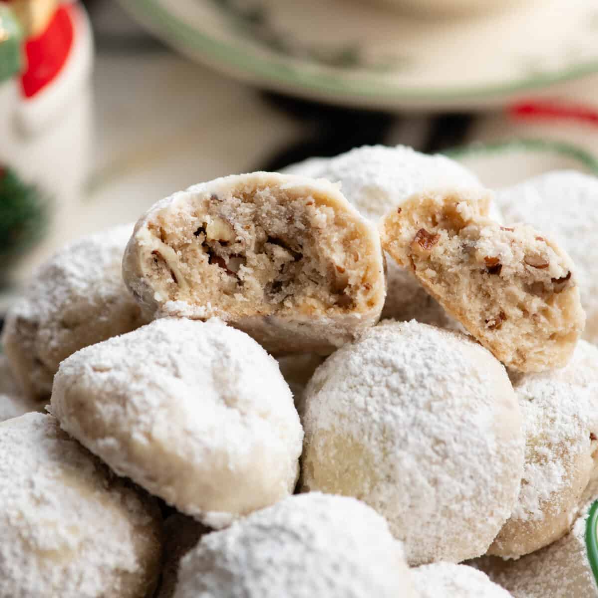 Pecan Snowball Cookies are easy to make with 7 ingredients. They are buttery, nutty, and melt-in your mouth delicious. These butterball cookies are an old family recipe, and a must-make Christmas cookie! snowball cookies piled on a Christmas tree shaped plate, one cookie has been broken in half and both halves are facing the camera so you can see the moist texture with pecans inside. There is a Christmas snowman and tea cup and saucer in the background