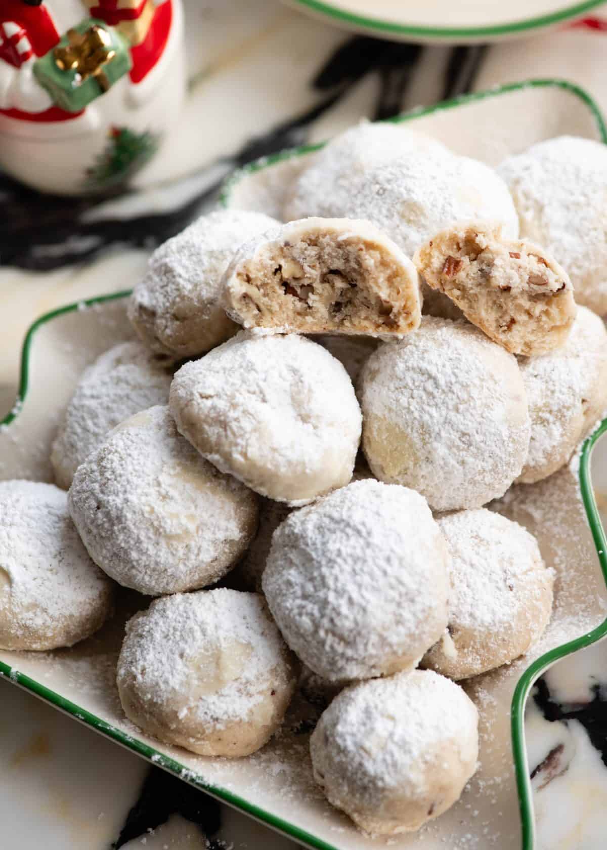snowball cookies piled on a Christmas tree shaped plate, one cookie has been broken in half and both halves are facing the camera so you can see the moist texture with pecans inside. There is a Christmas snowman and tea cup and saucer in the background