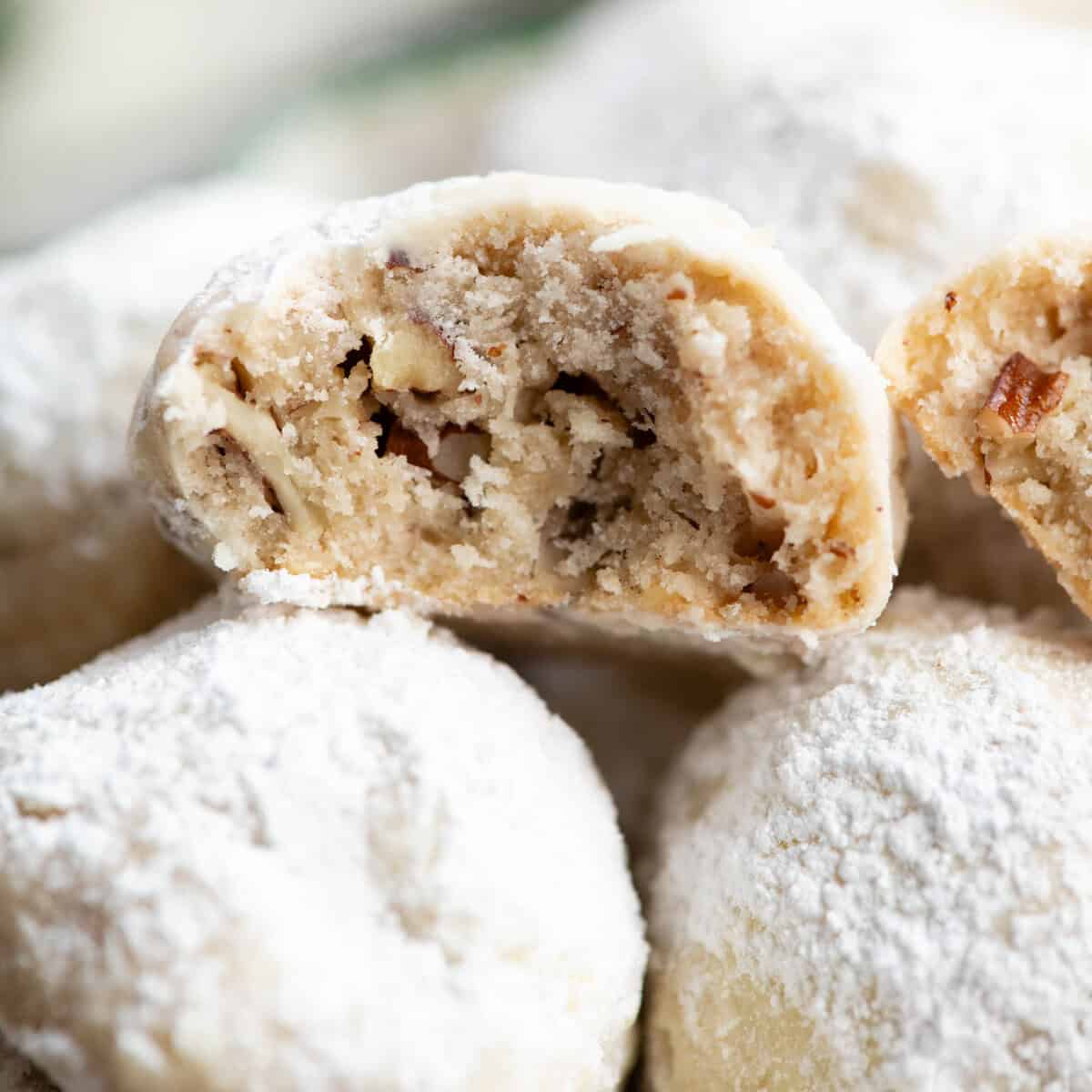 Pecan Snowball Cookies are easy to make with 7 ingredients. They are buttery, nutty, and melt-in your mouth delicious. These butterball cookies are an old family recipe, and a must-make Christmas cookie! close up photo of half of a snowball cookie with a bite taken out of it so the moist texture studded with pecans is visible. It's sitting on top of a pile of more snowball cookies and another half of the cookie is resting to the right side of it.