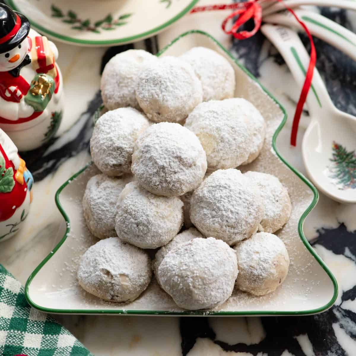 snowball cookies piled on a Christmas tree shaped plate with a Christmas snowman, Christmas measuring spoons and tea cup and saucer in the background