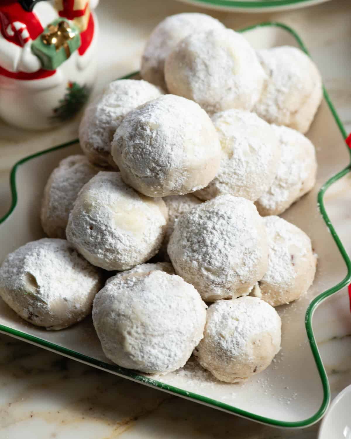 Pecan Snowball Cookies are easy to make with 7 ingredients. They are buttery, nutty, and melt-in your mouth delicious. These butterball cookies are an old family recipe, and a must-make Christmas cookie! snowball cookies piled on a Christmas tree shaped plate with a Christmas snowman and tea cup and saucer in the background
