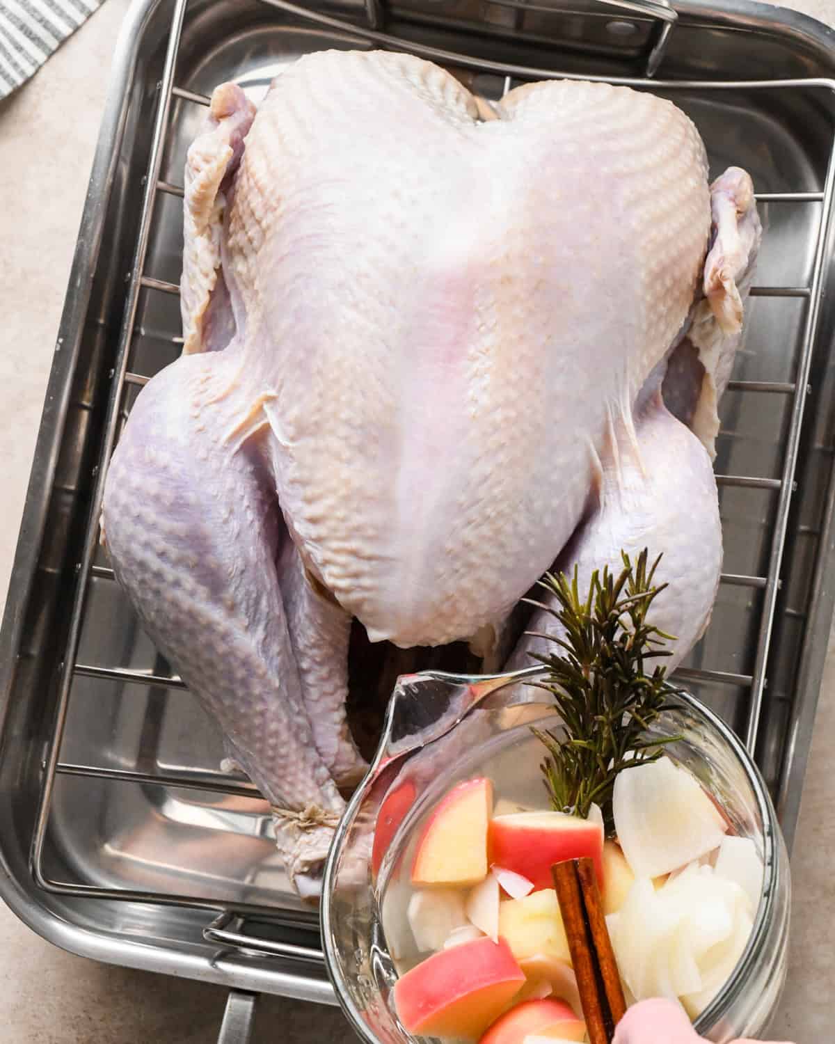 aromatics being poured into the cavity of a whole, uncooked turkey on a wire rack in a roasting pan. 