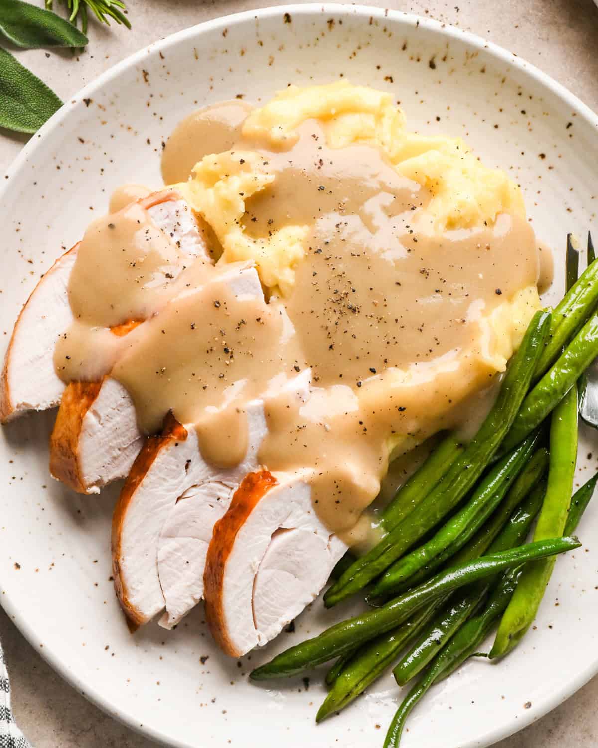 oven roasted turkey on a plate with mashed potatoes and green beans, all of it covered with gravy and cracked black pepper