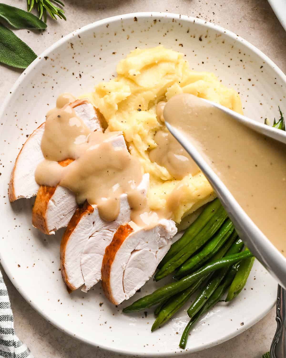 gravy being poured over oven roasted turkey on a plate with green beans and mashed potatoes. 