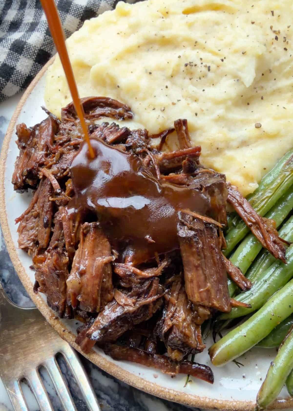 gravy being poured over slow cooker pot roast shredded on a white ceramic plate with mashed potatoes and green beans also on the plate.