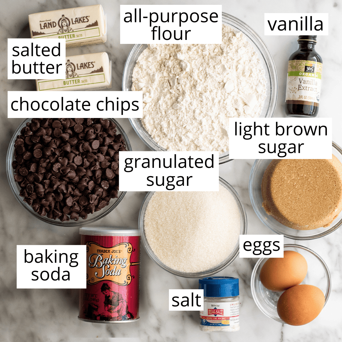 This chocolate chip cookie cake recipe is soft, chewy, slightly doughy and decorated with the best chocolate buttercream frosting. It's easy to make, large enough to feed a crowd, and even better than store-bought! overhead photo of the ingredients in this chocolate chip cookie cake recipe in bowls and labeled like flour, sugar, brown sugar, eggs, salt, baking soda, vanilla, butter and chocolate chips