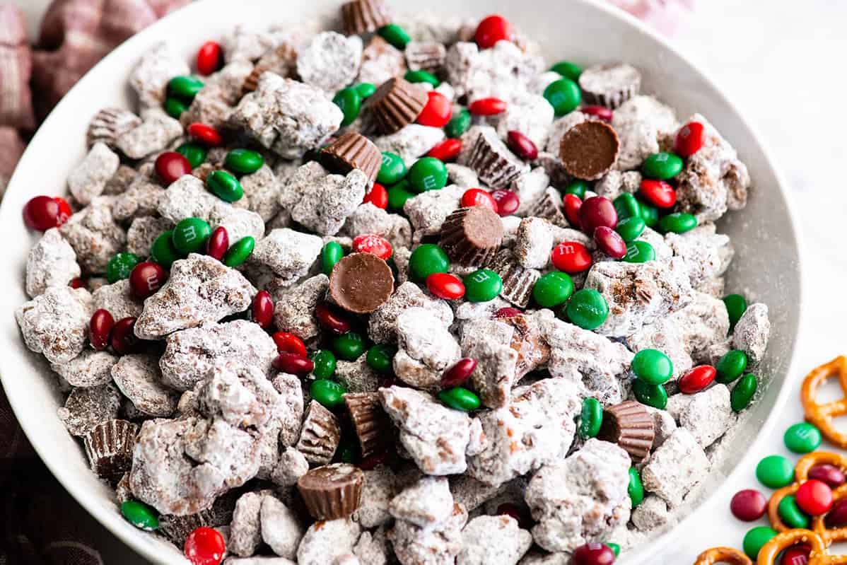 a bowl of Reindeer Food in a white serving bowl up - cereal thickly coated in peanut butter and chocolate and powdered sugar, there's also mini peanut butter cups and read and green Christmas M&Ms. There are pretzels and M&Ms in the bottom right corner and a red plaid towel in the top right corner.