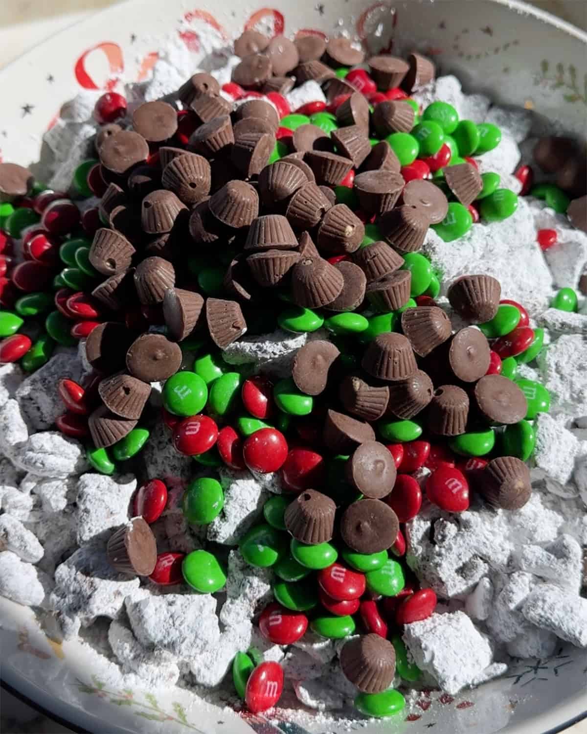 making reindeer food - mini peanut butter cups and christmas M&Ms added to the reindeer chow mixture in a christmas bowl before mixing