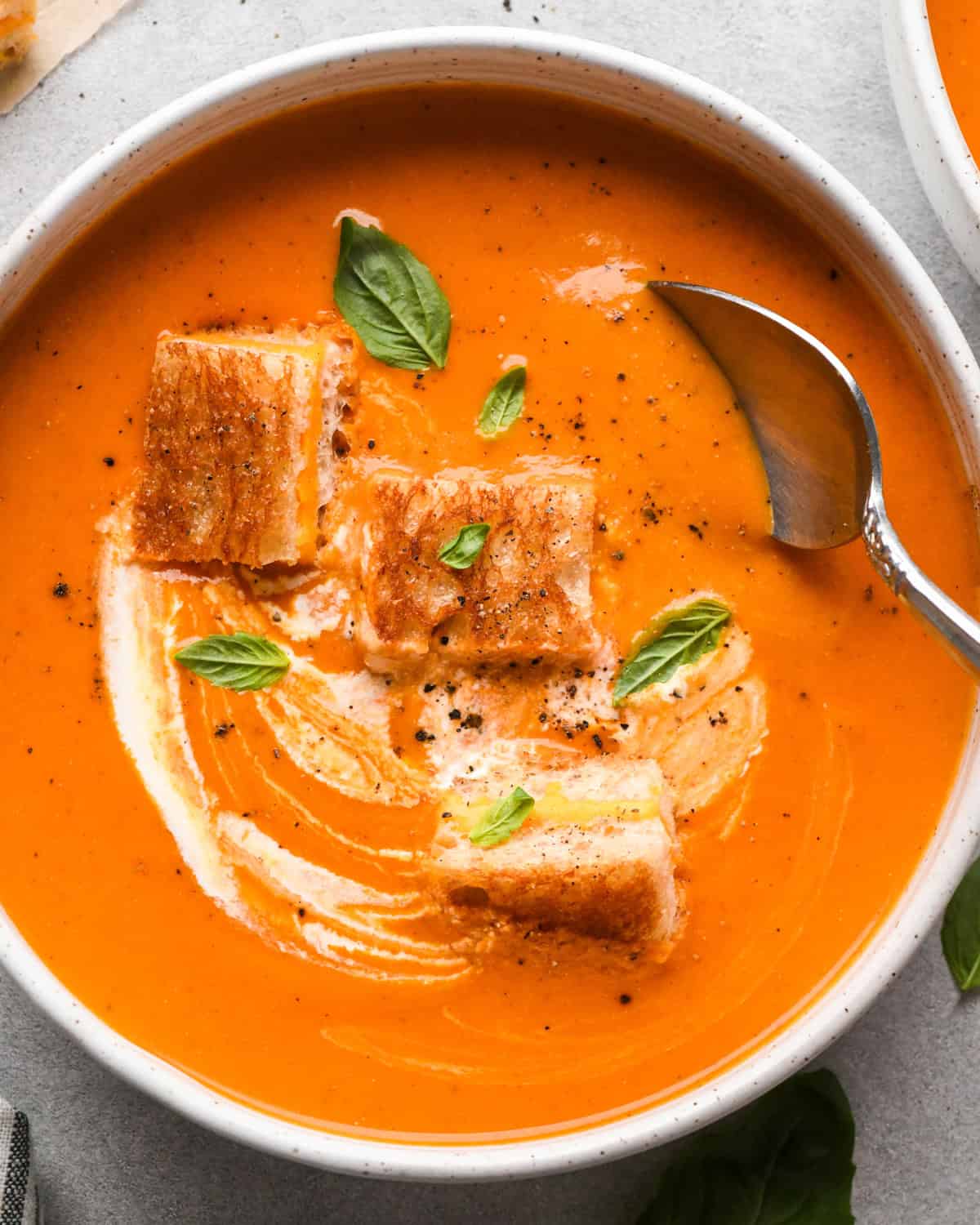 A bowl of creamy tomato soup with a spoon taking a scoop, and a swirl of cream on top. Three toasted grilled cheese croutons and fresh basil leaves garnish the tomato soup. The edge of a striped napkin is in the lower left hand corner 