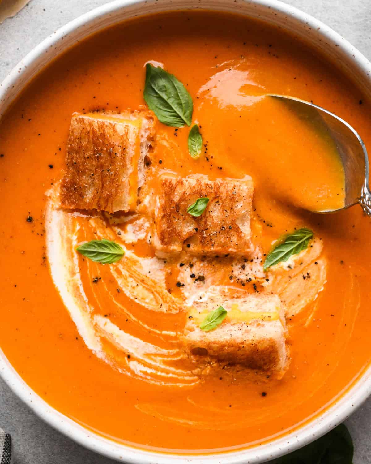 A bowl of creamy tomato soup with a spoon taking a scoop, and a swirl of cream on top. Three toasted grilled cheese croutons and fresh basil leaves garnish the tomato soup. The edge of a striped napkin is in the lower left hand corner 