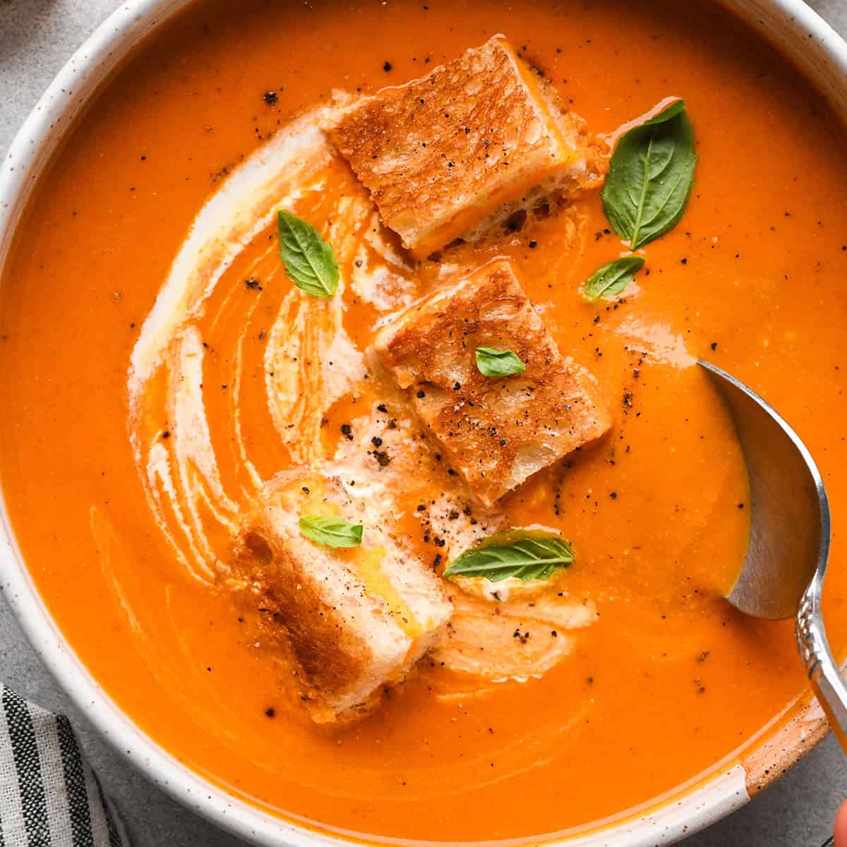 A bowl of creamy tomato soup with a spoon taking a scoop, and a swirl of cream on top. Three toasted grilled cheese croutons and fresh basil leaves garnish the tomato soup. The edge of a striped napkin is in the lower left hand corner 