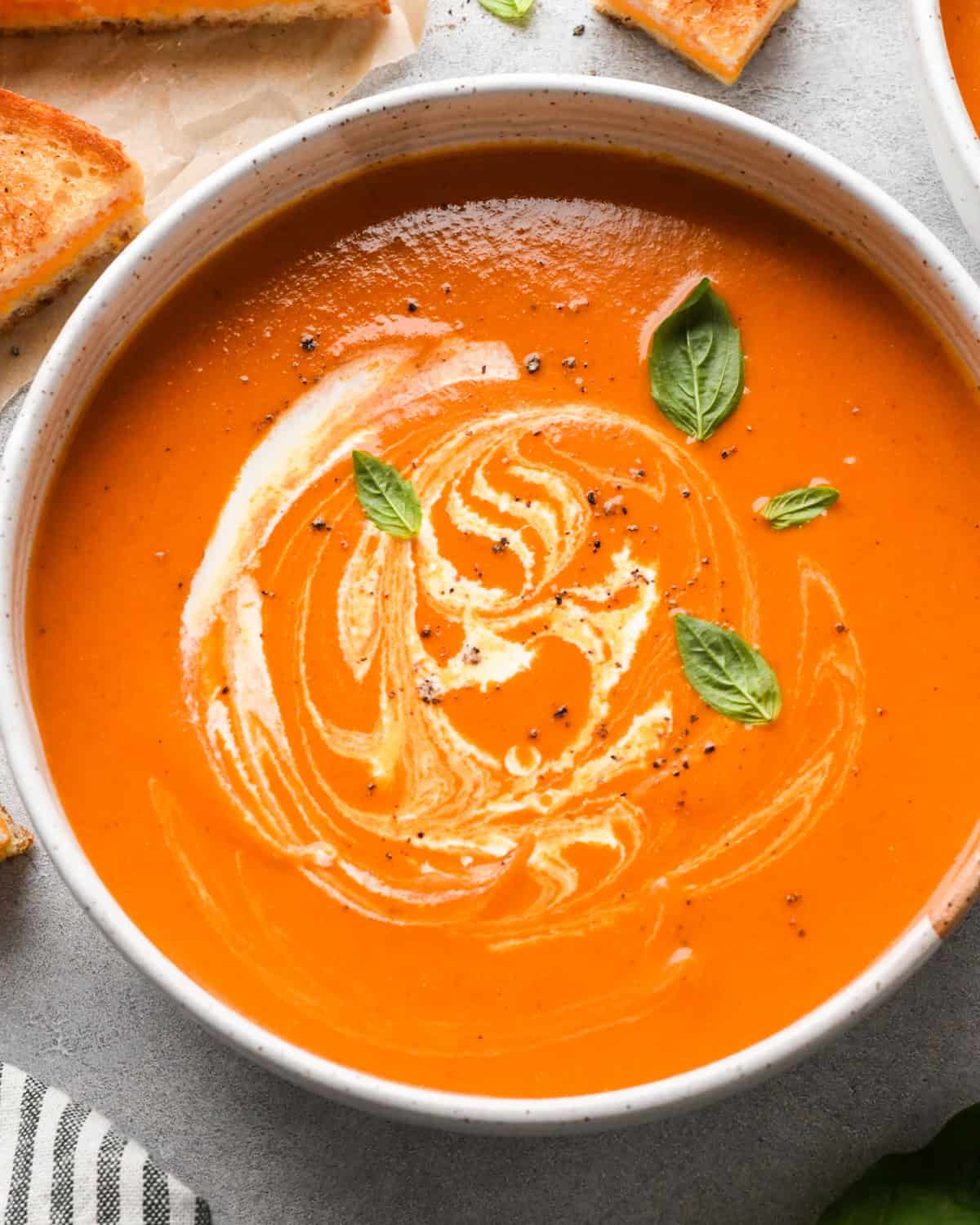 a bowl of tomato soup garnished with a swirl of heavy cream, black pepper and basil leaves. There are grilled cheese sandwiches in the top left corner 