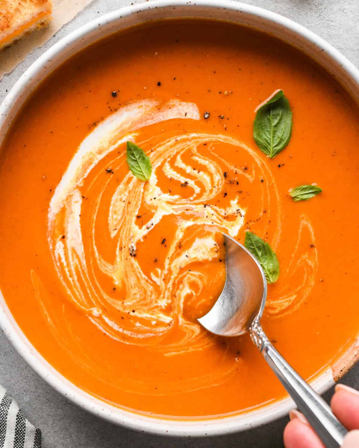 a bowl of tomato soup with a swirl of cream on top garnished with black pepper and basil leaves. A hand holding a spoon is taking a bite of the soup . 