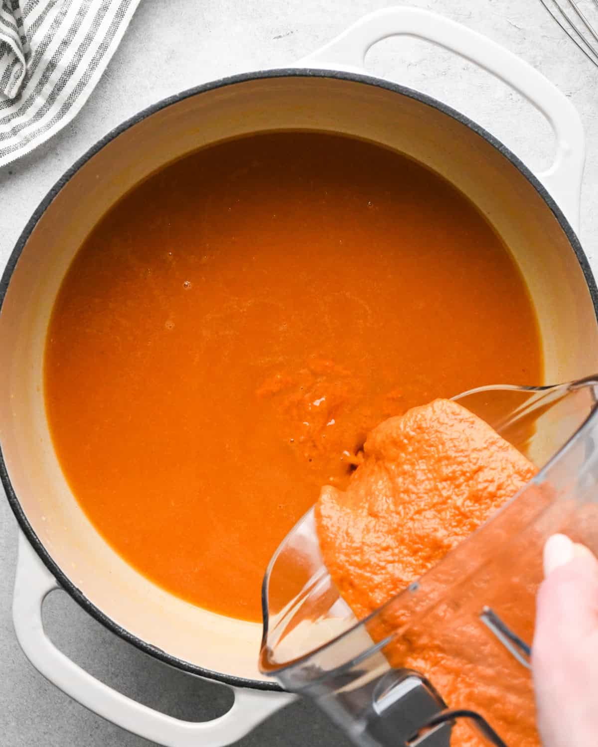 tomato puree being poured from a blending container into a white pot with vegetable broth in it making tomato soup