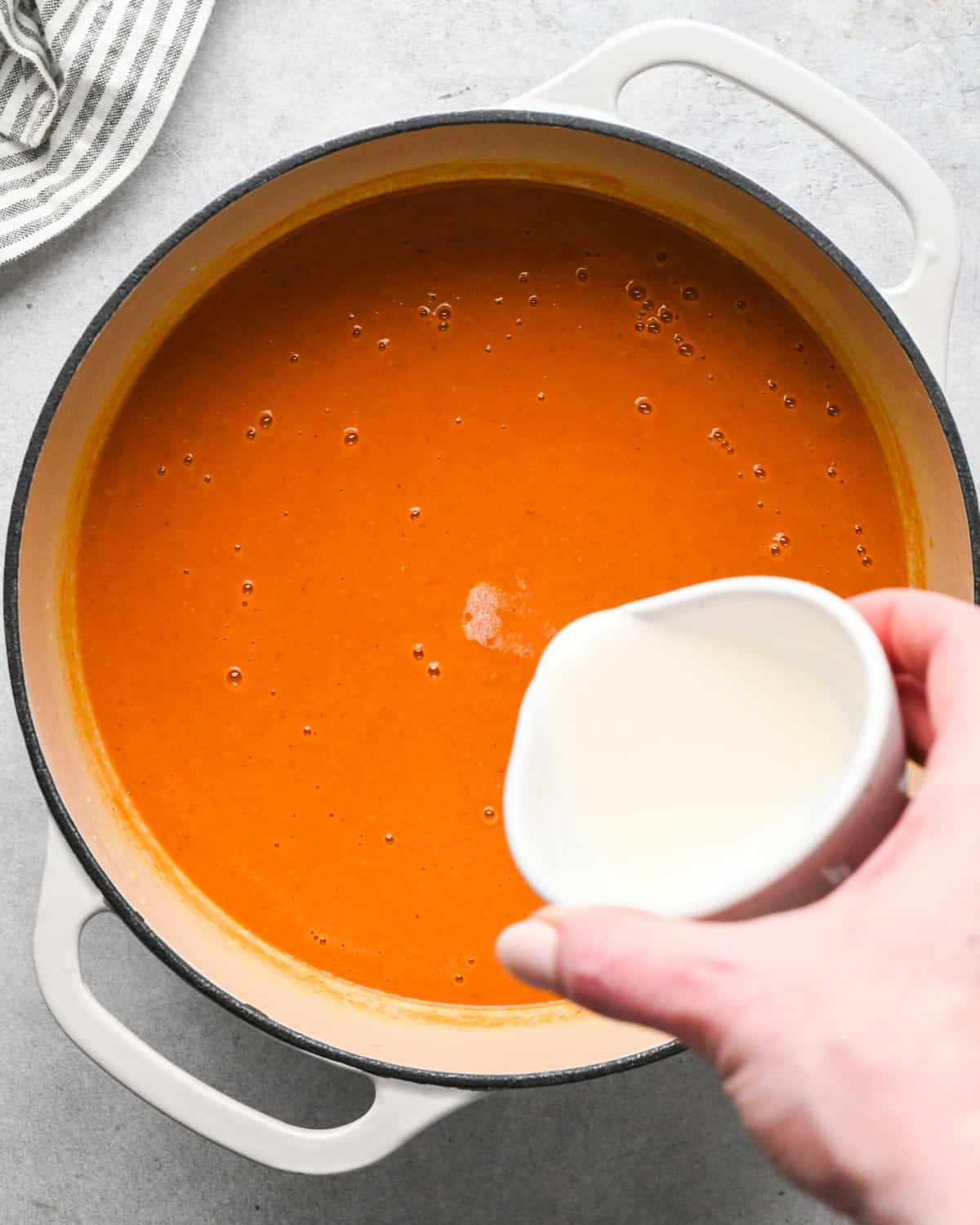 a hand pouring heavy cream into a pot of tomato soup 