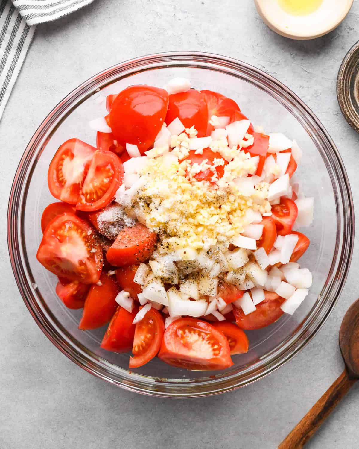 A glass bowl with chopped tomatoes, diced onions, olive oil, crushed garlic, and a sprinkle of black pepper— in the process of making tomato soup. A wooden spoon lies beside the bowl on a gray countertop, with a striped cloth and small bowl of olive oil partially visible.