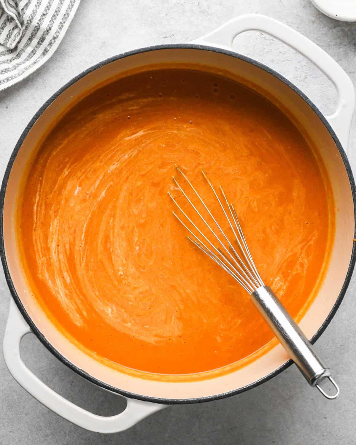 a pot of tomato soup with the heavy cream partially whisked into it, the whisk is in the pot and a striped towel sits beside the pot in the top left corner 