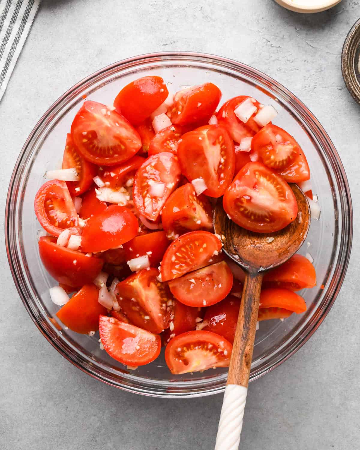 A glass bowl with chopped tomatoes and diced onions is mixed with a wooden spoon, the start to making this tomato soup recipe. 