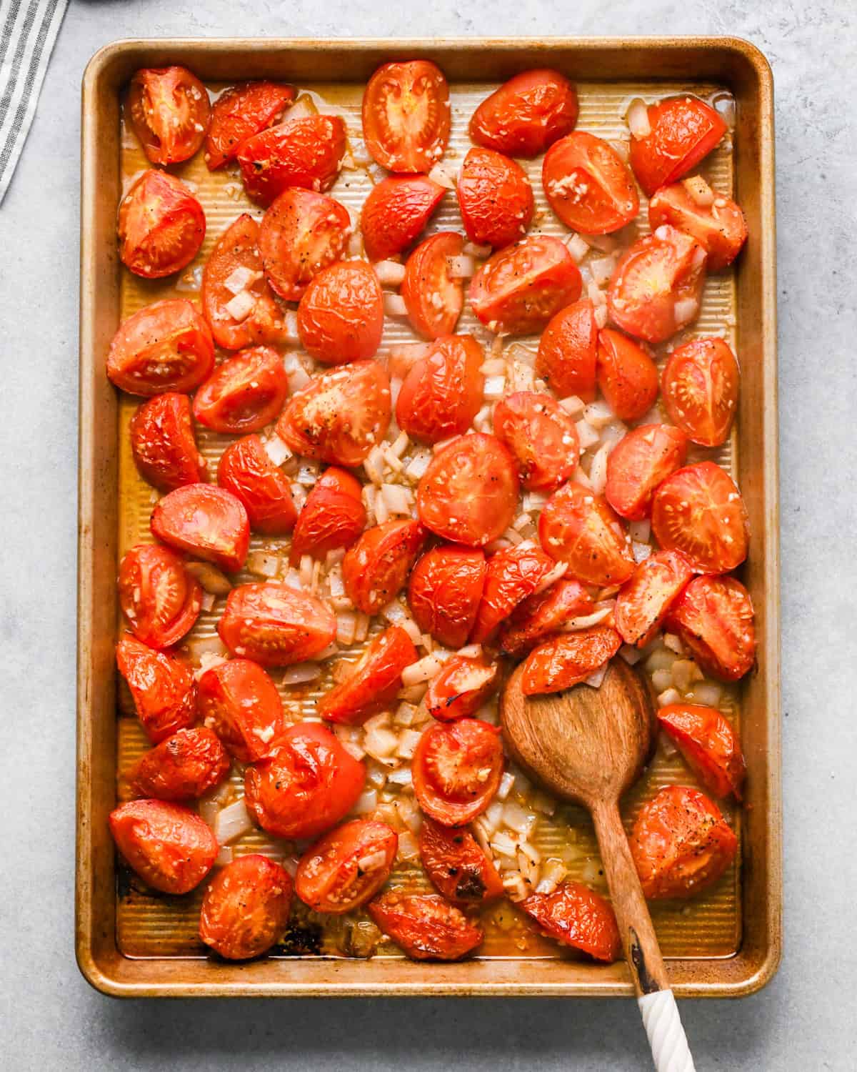 tomatoes, garlic, olive oil and onions being stirred with a wooden spoon on a baking sheet after roasting for 15 minutes making this homemade tomato soup recipe