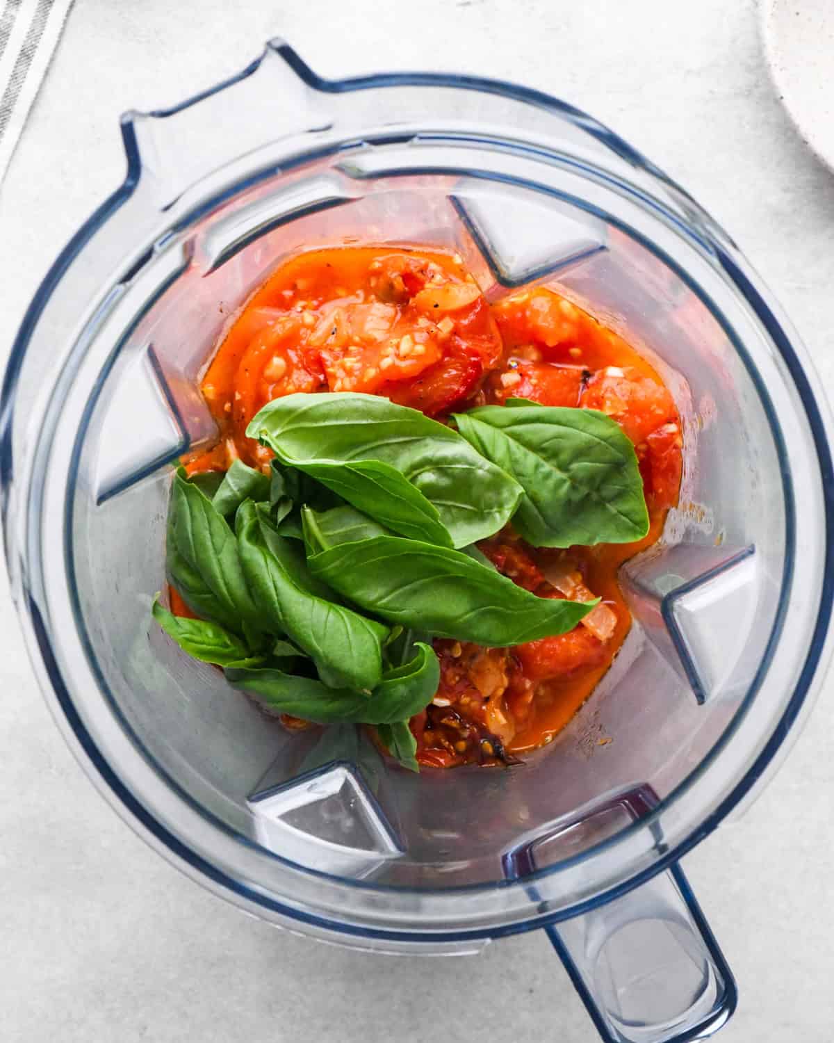 fresh basil leaves added on top of a roasted tomato mixture in the container of a vitamix blender before blending making tomato soup