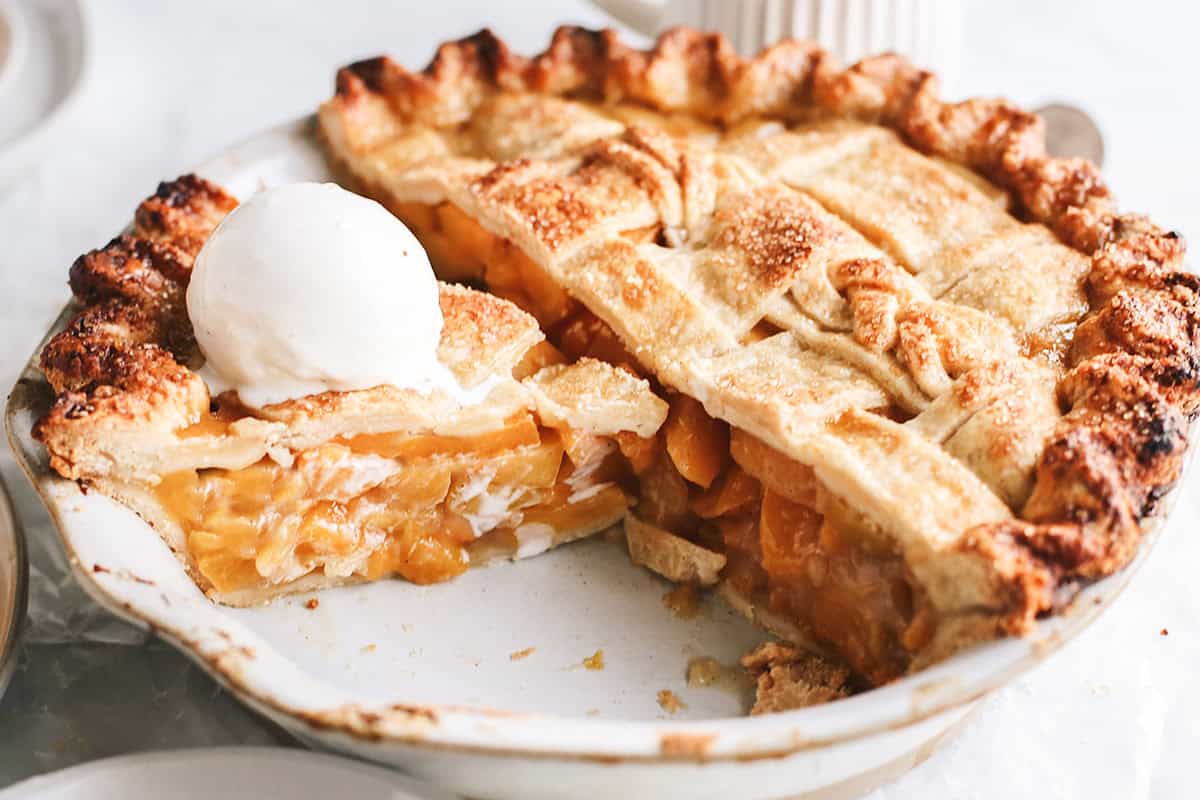 front view of a peach pie in a pie dish with a slice cut out with ice cream on top