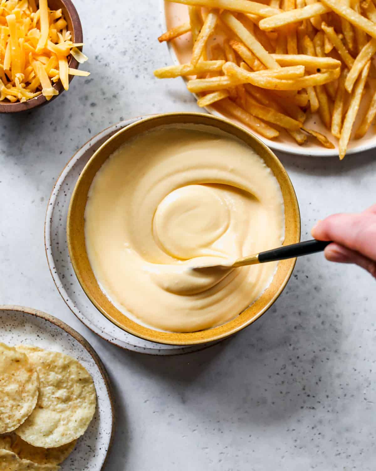 overhead view of a hand stirring cheese sauce in a bowl with fries and chips on plates next to it