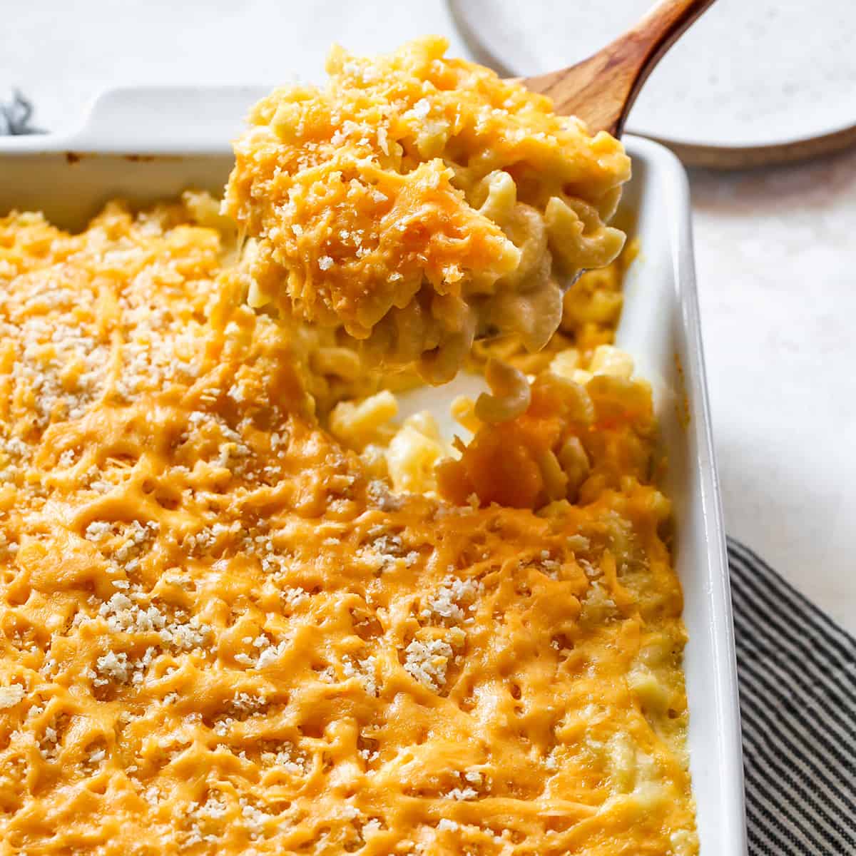 front view of a wooden spoon taking a scoop of Baked Mac and Cheese out of a white baking dish