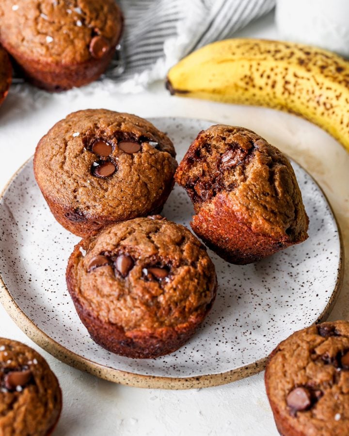three Healthy Banana Chocolate Chip Muffins on a plate, one with a bite taken out of it. 