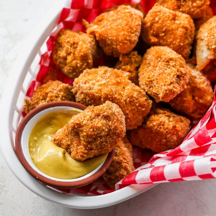 Homemade Chicken Nuggets in a basket with a red and white checkered liner, one being dipped into a bowl of mustard. 
