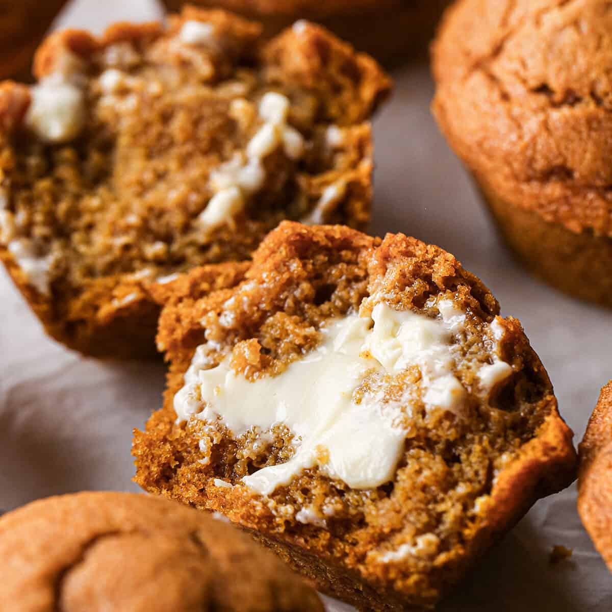 half of a pumpkin muffin with butter on it. the other half of the muffin and another full muffin is in the background.