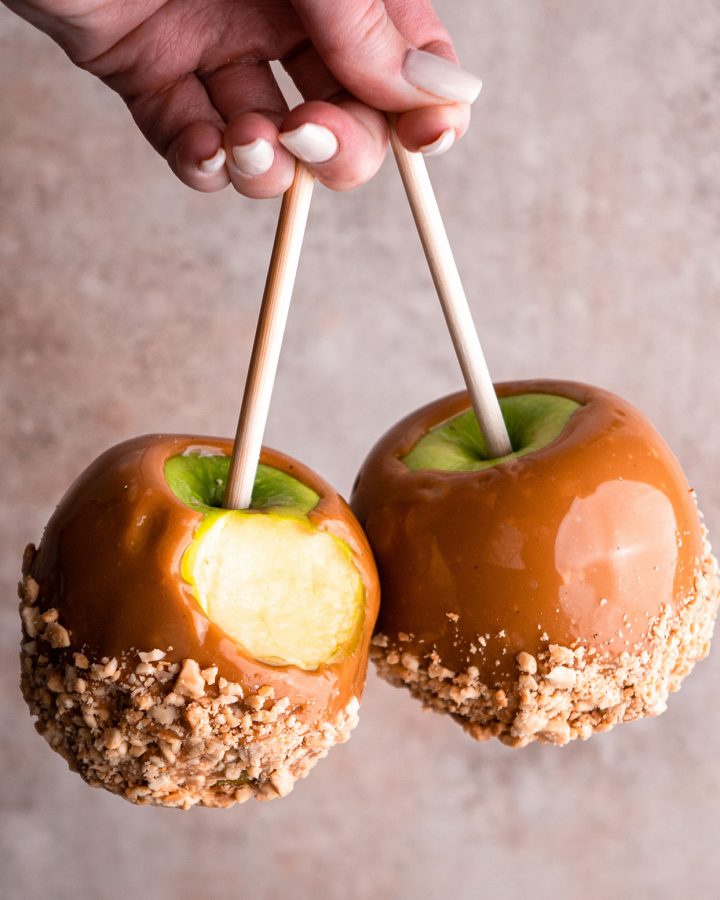 a hand holding two Homemade Caramel Apples, one with a bite taken out of it 