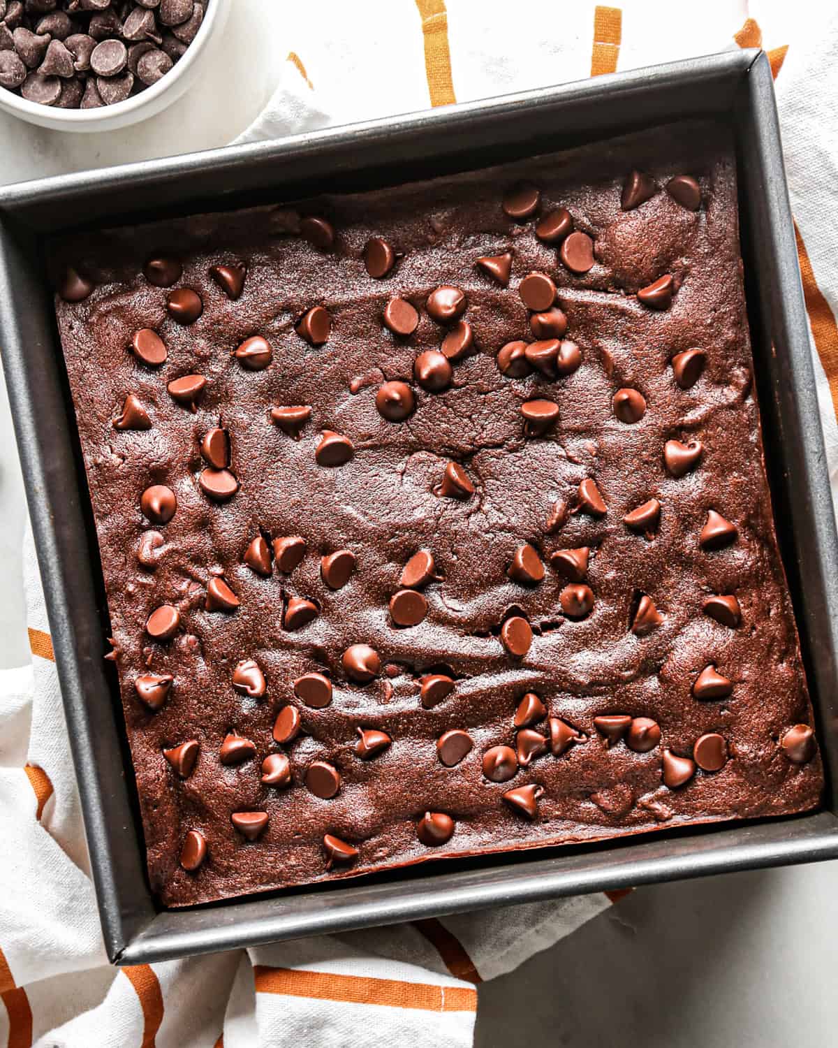 baked pumpkin brownies in a square metal baking pan on top of a white and orange striped towel with a bowl of chocolate chips nearby
