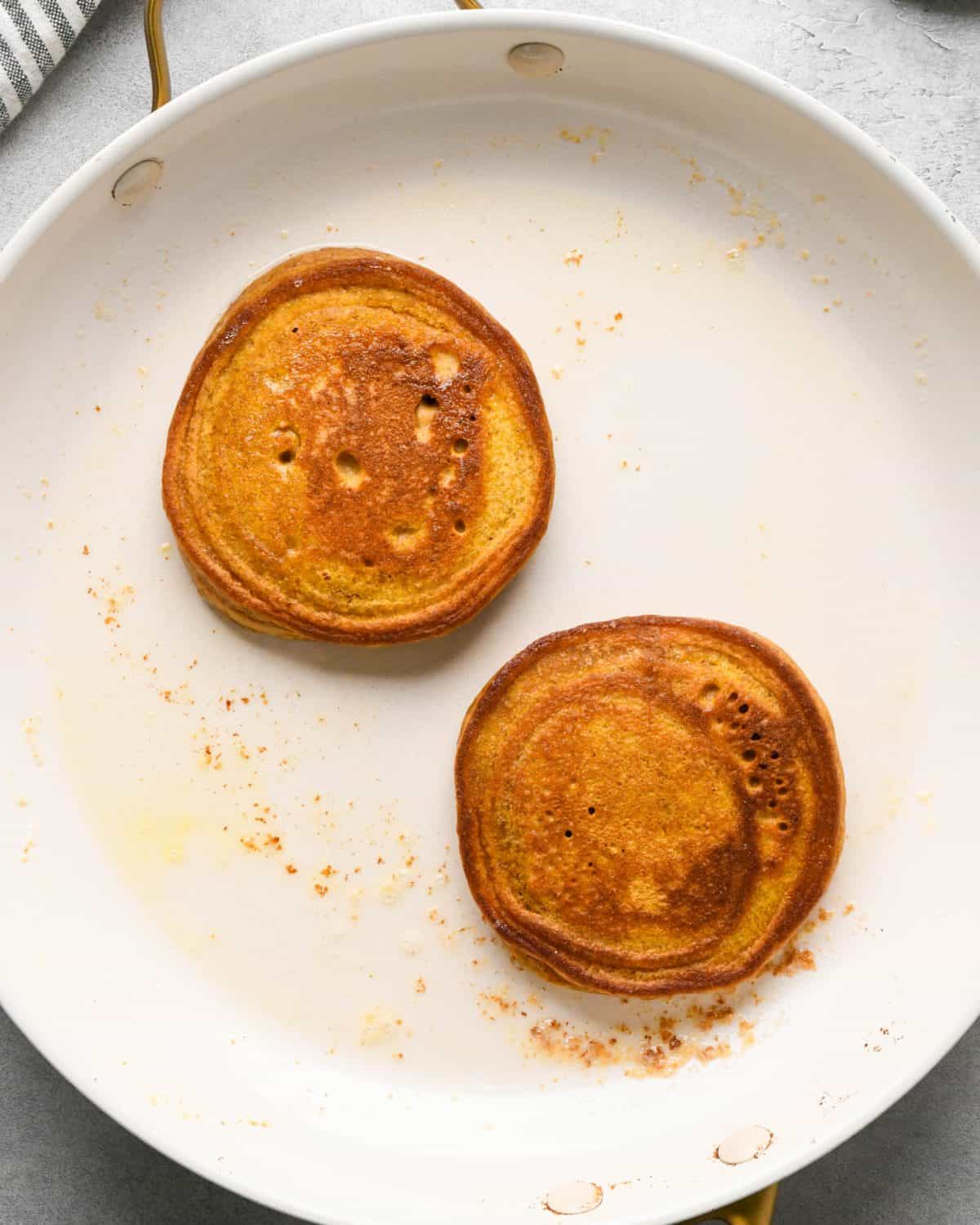 two pumpkin pancakes cooking on the second side in melted butter in a white nonstick frying pan.