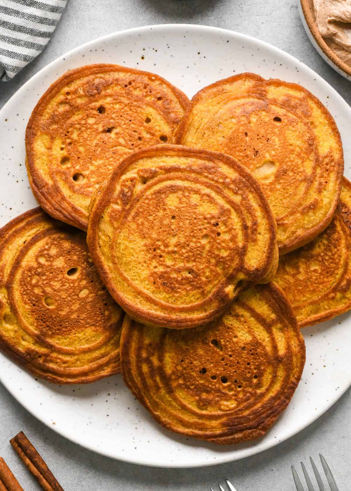 6 pumpkin pancakes on a white ceramic plate surrounded by a bowl of cinnamon butter, cinnamon sticks, and forks.