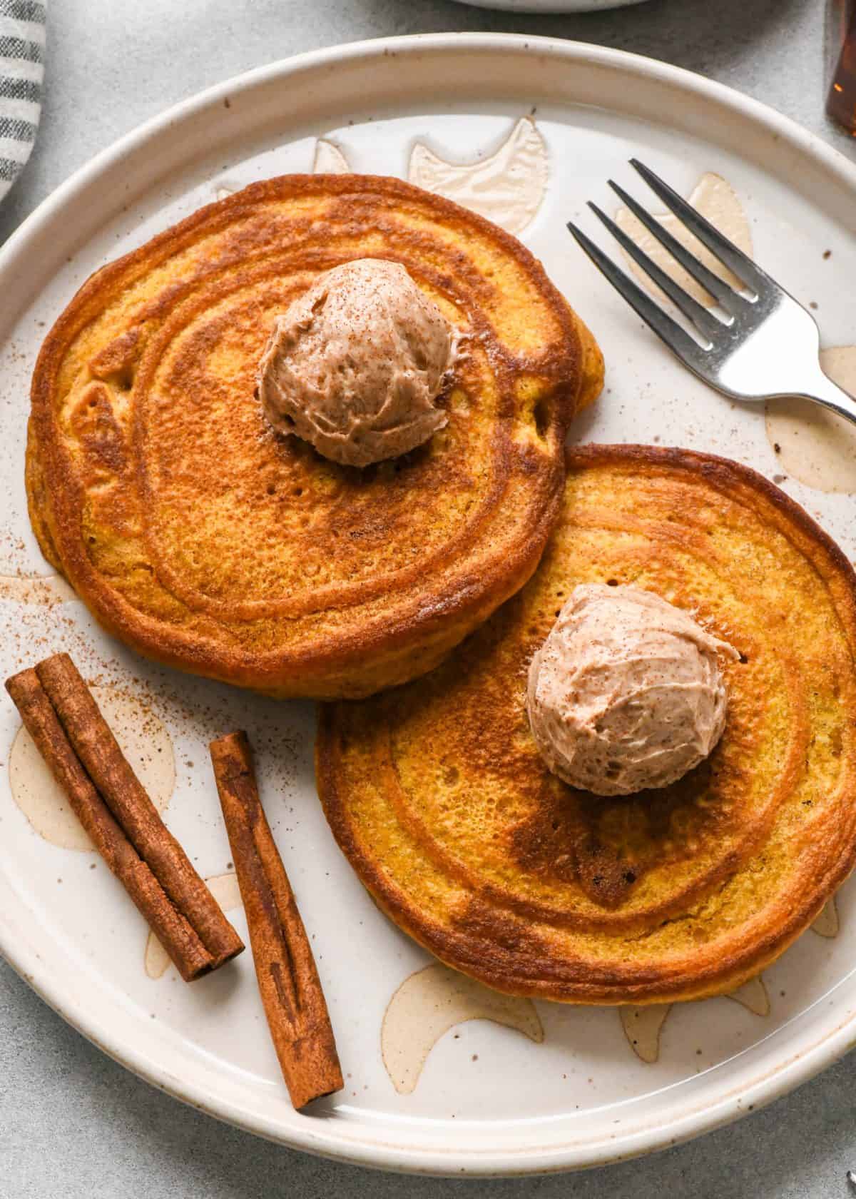 two pumpkin pancakes on a white ceramic plate, each has a scoop of cinnamon butter on top. There's a fork and two cinnamon stick son the plate too.